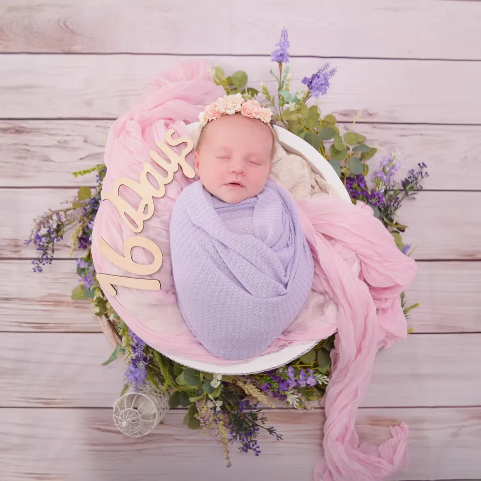 Newborn swaddled in lilac in a white bowl beside a script sign reading sixteen days