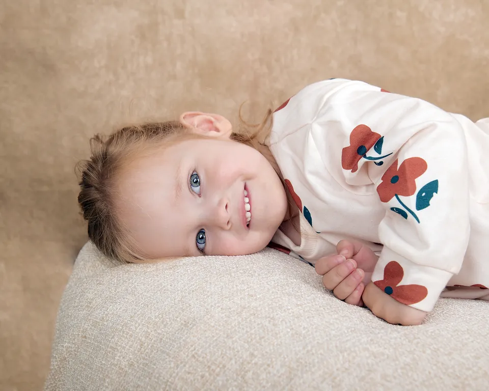 Young child in a poppy-print top resting on a cream cushion, smiling upward