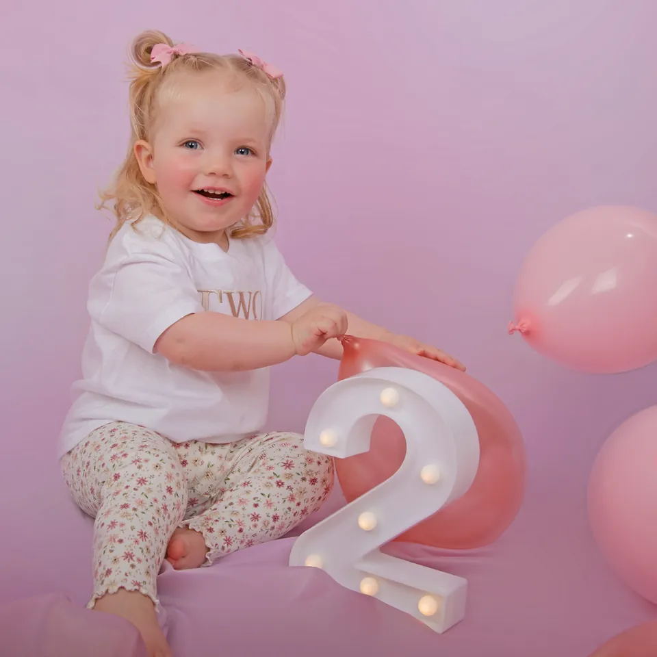 Toddler sitting beside a light-up number two and pink balloons at a birthday shoot