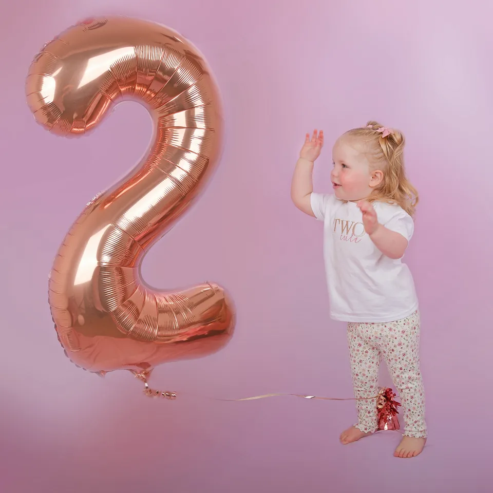Toddler waving beside a giant rose-gold number two balloon on pink