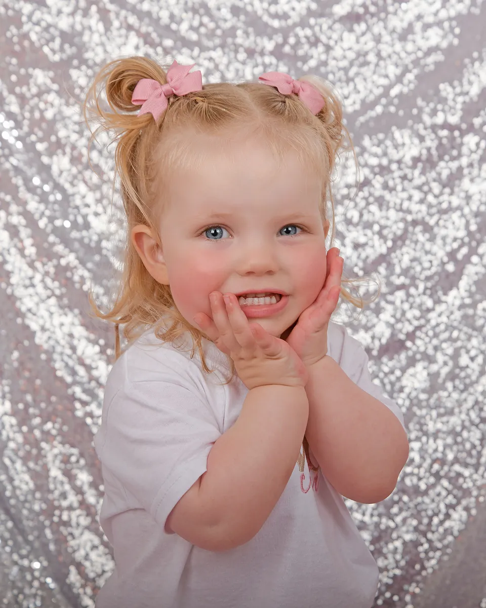 Toddler with pigtails, hands pressed to her cheeks, against a silver sequin backdrop