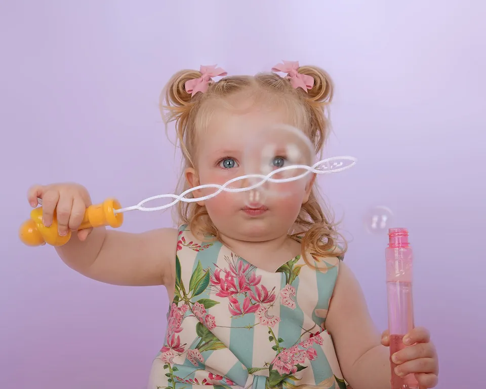 Toddler in a floral dress blowing bubbles from a yellow wand on lilac