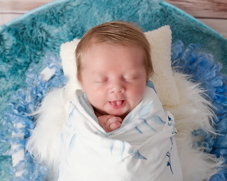 Closer landscape crop of a sleeping newborn swaddled in white in a teal fur nest