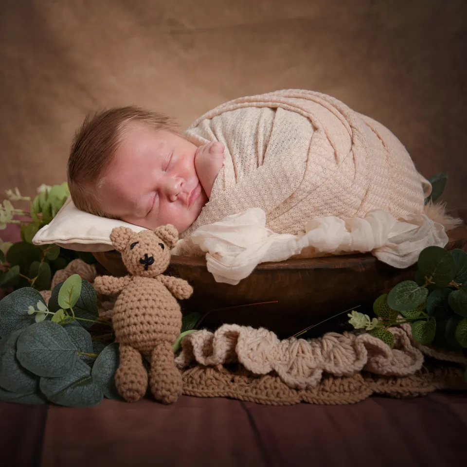 Newborn asleep in a wooden bowl with a crocheted teddy and eucalyptus
