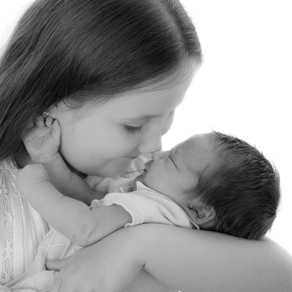 Black-and-white portrait of an older child leaning cheek to cheek with a newborn