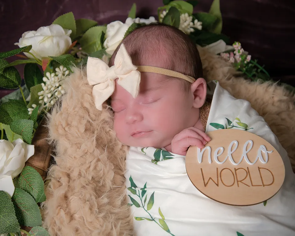Newborn asleep in a floral nest holding a wooden disc reading hello world