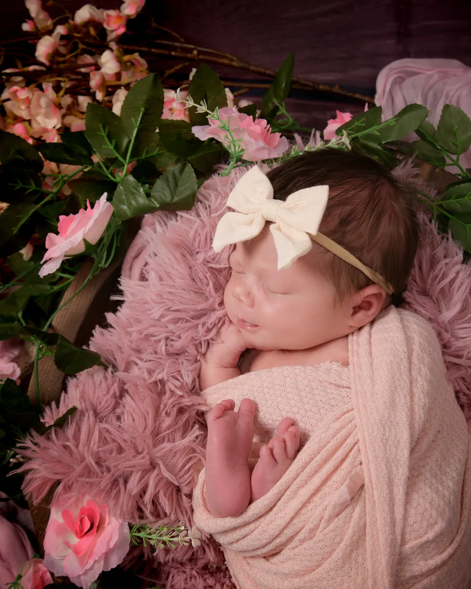 Newborn in a pink wrap asleep on faux fur amid pink blossom and foliage