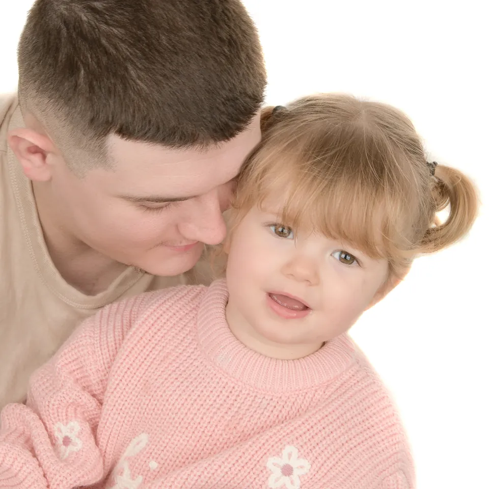 Toddler in a pink jumper leaning against an adult's shoulder on a white backdrop